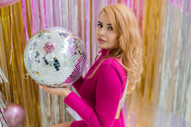 Mujer rubia glamour en vestido rosa de lujo posando con bola de discoteca