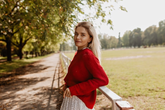 Mujer rubia gloriosa de pie relajado en el parque. Hermosa chica disfrutando de una bonita vista otoñal.