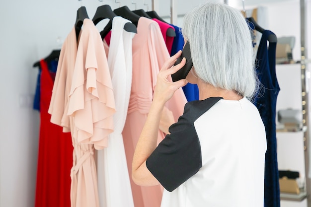 Mujer rubia hablando por teléfono móvil mientras elige ropa y busca vestidos en el estante en la tienda de moda. Vista trasera. Cliente boutique o concepto minorista