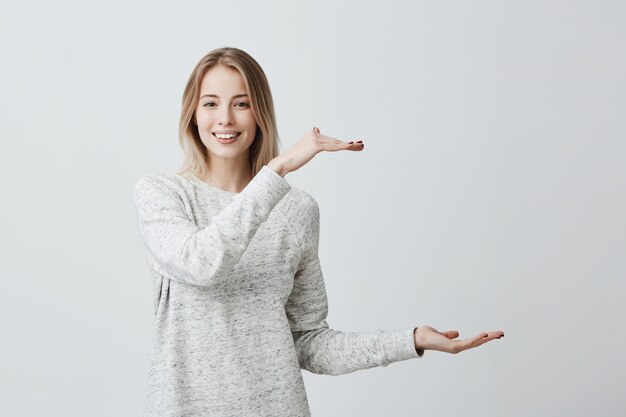 Mujer rubia hermosa emocionada en ropa casual que muestra con las manos longitud de la caja. La mujer contenta que usa un suéter suelto sonríe alegremente demuestra el tamaño de algo grande.