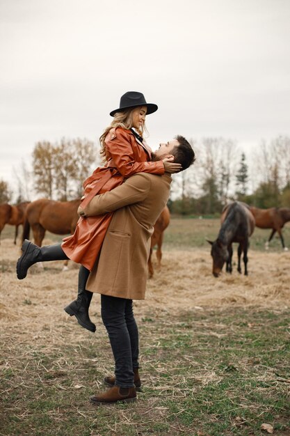 Mujer rubia y hombre moreno de pie en el campo con caballos marrones. Mujer con vestido negro, abrigo de cuero y sombrero y hombre con abrigo marrón. Hombre y mujer abrazándose.