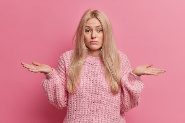 Una mujer rubia incierta extiende las palmas de las manos y duda que el interior no pueda elegir entre dos opciones viste un suéter cálido tejido