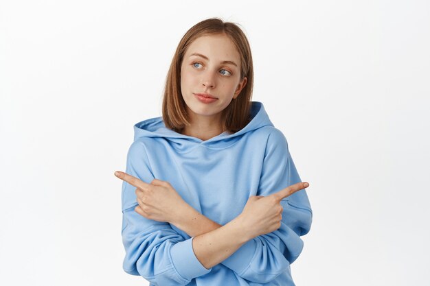 Mujer rubia indecisa hace su elección entre dos variantes, apuntando hacia los lados pero mirando a la izquierda, pensando, eligiendo artículos en oferta, pared blanca