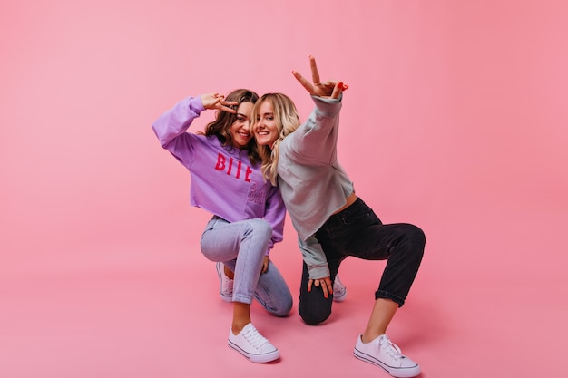 Mujer rubia inspirada en pantalón negro posando con el signo de la paz. Jocund chica morena pasar tiempo con su hermana.