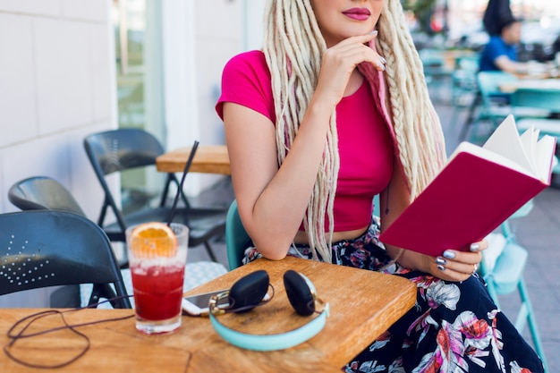 Mujer rubia inusual con rastas sentado en un café en la calle, sosteniendo el cuaderno, disfrutando del tiempo libre. Usar pantalones brillantes con estampado tropical.