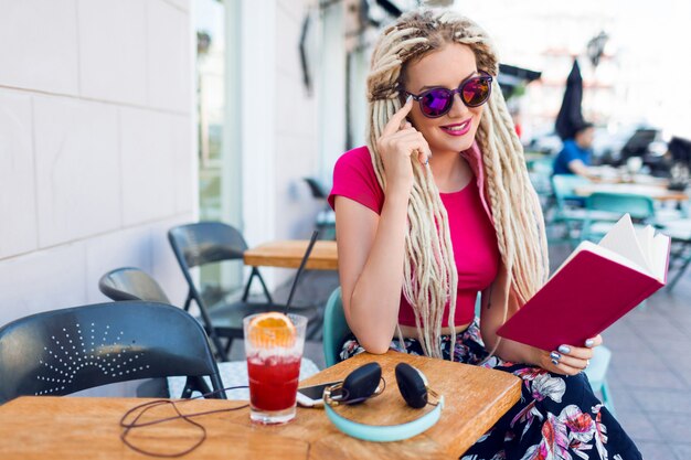 Mujer rubia inusual con rastas sentado en un café en la calle, sosteniendo el cuaderno, disfrutando del tiempo libre. Usar pantalones brillantes con estampado tropical.