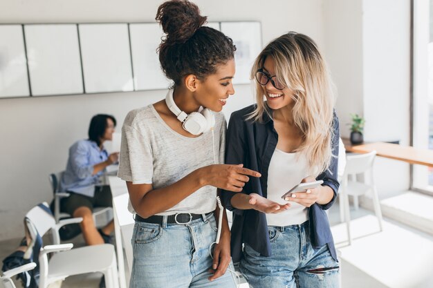 Mujer rubia en jeans y gafas con smartphone mientras habla con un amigo africano en la oficina. Estudiante bastante internacional en auriculares pasar tiempo con compañeros de la universidad.