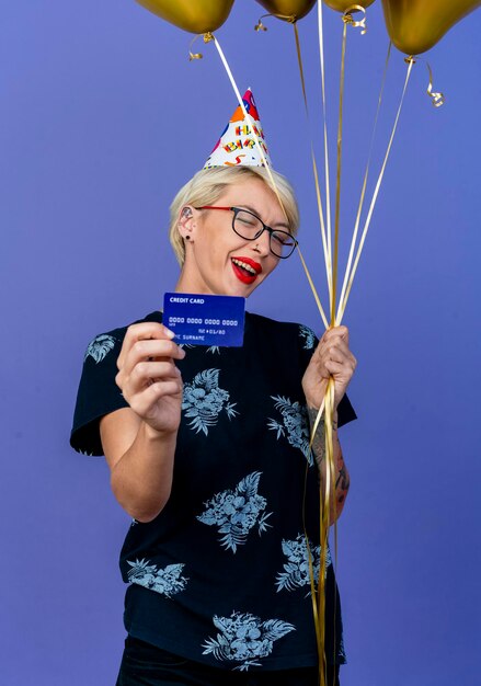Mujer rubia joven alegre del partido con gafas y gorra de cumpleaños sosteniendo globos y tarjeta de crédito guiñando un ojo en el frente aislado en la pared púrpura