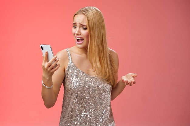 La mujer rubia joven arrogante preocupada preocupada quejándose gritando que el teléfono inteligente no puede llamar a un amigo sin señal que sostiene la pantalla del teléfono inteligente mira el móvil cabreado de mal humor argumentando, fondo rojo.