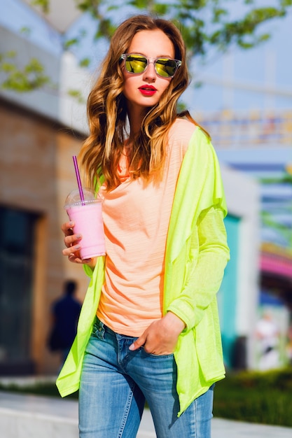 Mujer rubia joven inconformista con estilo en gafas de sol con cóctel posando al aire libre