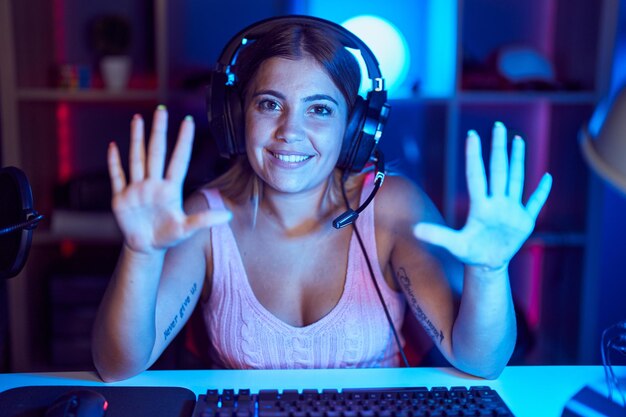 Mujer rubia joven jugando videojuegos con auriculares mostrando y apuntando con los dedos número diez mientras sonríe segura y feliz