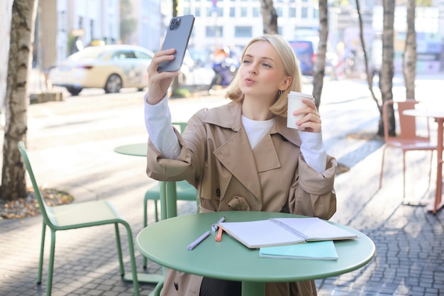 Mujer rubia joven de moda tomando una selfie con su bebida favorita en la cafetería pliegues labios