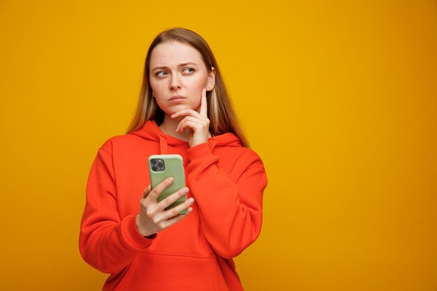 Mujer rubia joven pensativa manteniendo la mano en la barbilla sosteniendo y mirando el teléfono móvil