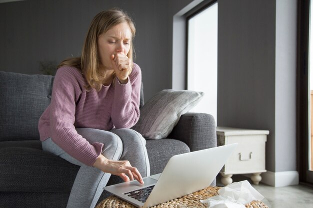 Mujer rubia joven que bosteza, escribiendo en la computadora portátil con una mano