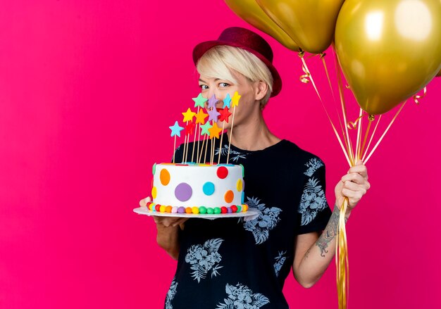 Mujer rubia joven sonriente del partido que lleva el sombrero del partido que sostiene los globos y el pastel de cumpleaños con las estrellas que miran al frente aislado en la pared rosada con el espacio de la copia