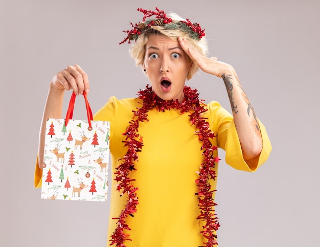 Mujer rubia joven sorprendida con corona de Navidad y guirnalda de oropel alrededor del cuello sosteniendo una bolsa de regalo de Navidad mirando a la cámara tocando la cabeza aislada sobre fondo blanco