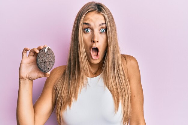 Mujer rubia joven sosteniendo piedra pómez asustada y asombrada con la boca abierta por cara de incredulidad sorpresa