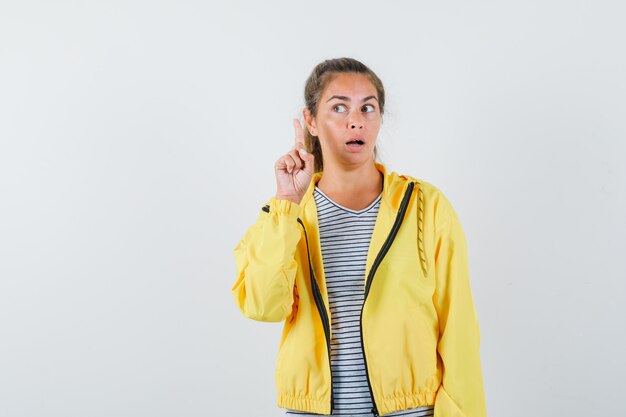 Mujer rubia levantando el dedo índice en gesto de eureka y mirándolo con chaqueta de bombardero amarilla y camisa a rayas y mirando serio