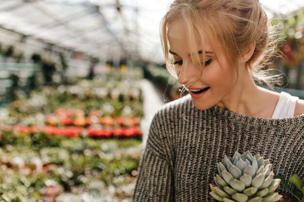 Mujer rubia con lunar encima de su labio sostiene suculentas. Mujer en suéter gris posando en la tienda de plantas.