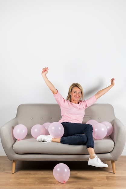 Mujer rubia madura emocionada que se sienta en el sofá con los globos rosados que aumentan sus brazos