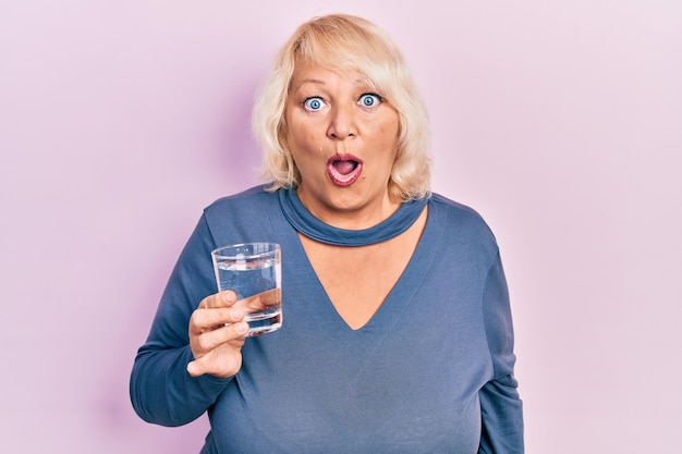 Foto gratuita mujer rubia de mediana edad bebiendo un vaso de agua asustada y asombrada con la boca abierta para la sorpresa cara de incredulidad