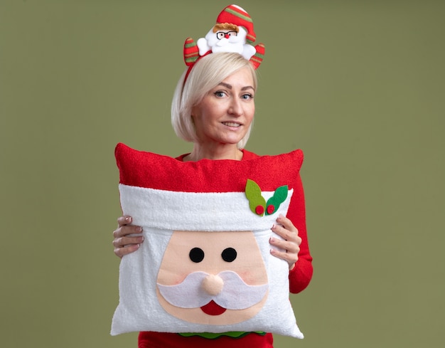 Mujer rubia de mediana edad complacida con diadema de santa claus y suéter de navidad con almohada de santa claus mirando a cámara mirando aislado sobre fondo verde oliva con espacio de copia