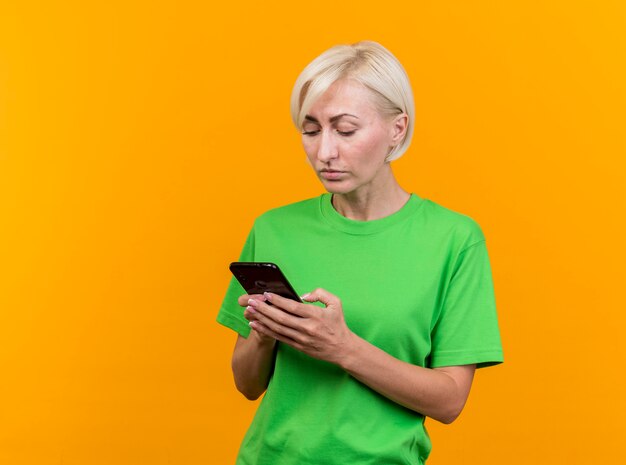 Mujer rubia de mediana edad concentrada usando su teléfono móvil aislado en la pared amarilla