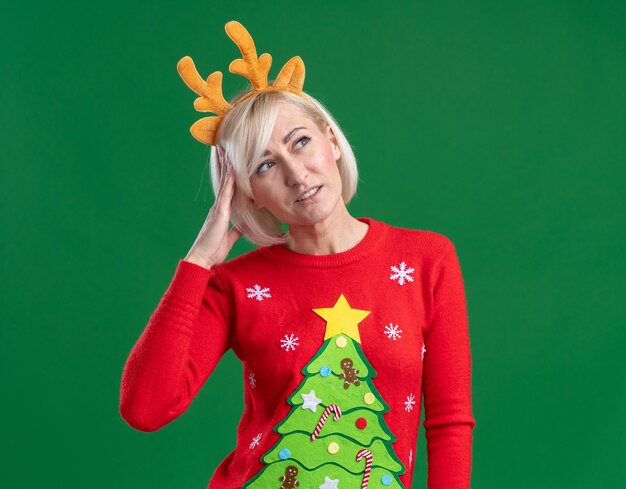 Mujer rubia de mediana edad confundida con diadema de cuernos de reno de Navidad y suéter de Navidad mirando al lado tocando la cabeza aislada sobre fondo verde con espacio de copia