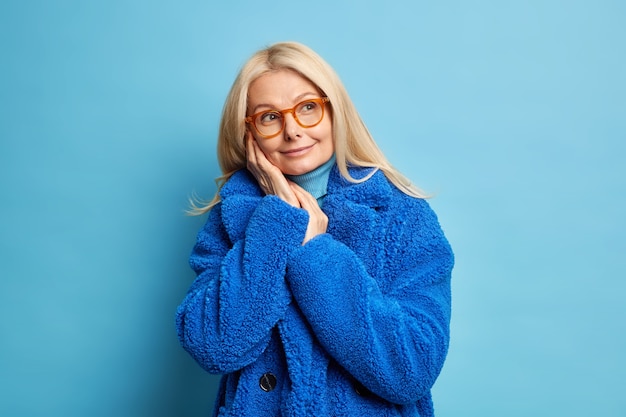 mujer rubia de mediana edad en gafas recuerda algo agradable mantiene las manos cerca de la cara piensa en el futuro mira a un lado sonríe suavemente viste abrigo de invierno azul cálido.