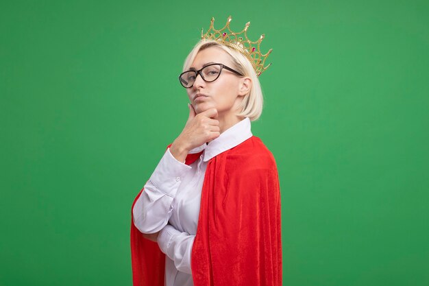 Mujer rubia de mediana edad segura de superhéroe en capa roja con gafas y corona de pie en la vista de perfil mirando al frente poniendo la mano en la barbilla aislada en la pared verde con espacio de copia