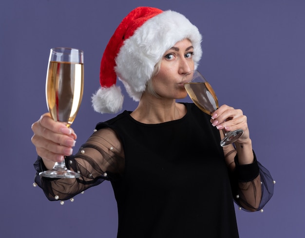 Foto gratuita mujer rubia de mediana edad con sombrero de navidad mirando a la cámara sosteniendo dos copas de champán bebiendo una y estirando otra hacia la cámara aislada sobre fondo púrpura