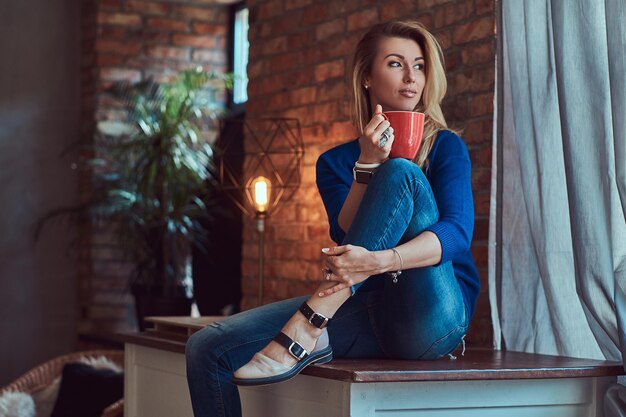 Mujer rubia de moda sostiene una taza de café mientras se sienta en una mesa contra una pared de ladrillos en un estudio con un interior de loft.