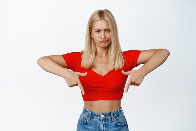 Foto gratuita una mujer rubia molesta haciendo una mueca señalando con el dedo algo malo se siente preocupada al mostrar un anuncio decepcionante de fondo blanco