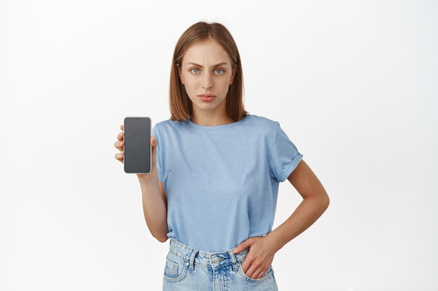Mujer rubia molesta que muestra la pantalla de su teléfono inteligente con la expresión de la cara decepcionada con el ceño fruncido. Mujer disgustada se queja, muestra la interfaz del teléfono móvil, pared blanca