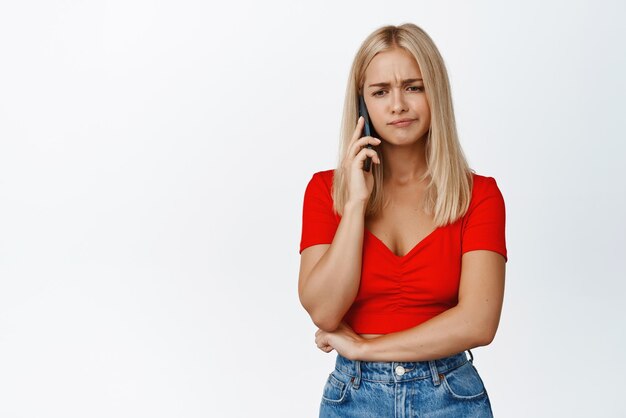 Una mujer rubia molesta recibe malas noticias por una llamada telefónica hablando por teléfono móvil con una expresión facial preocupada de fondo blanco