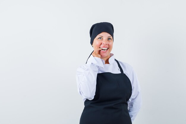 Mujer rubia mordiendo el dedo índice, sonriendo en uniforme de cocinero negro y luciendo bonita