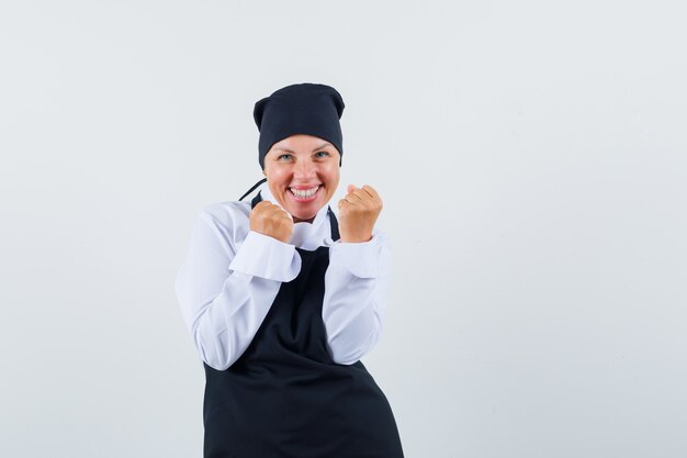 Mujer rubia mostrando gesto de éxito en uniforme de cocinero negro y mirando feliz. vista frontal.