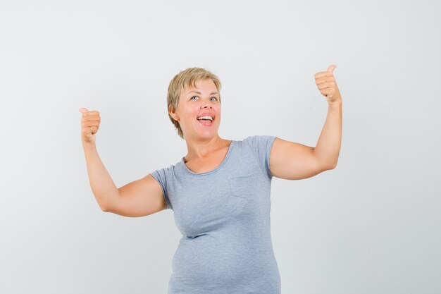 Mujer rubia mostrando gesto de ganador en camiseta azul claro y mirando alegre, vista frontal.