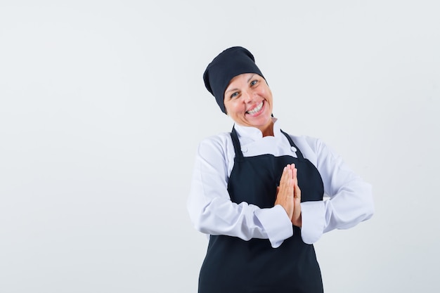 Mujer rubia mostrando gesto de namaste en uniforme de cocinero negro y luciendo bonita. vista frontal.