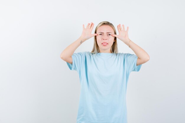 Mujer rubia mostrando gesto de orejas, sacando la lengua en camiseta azul y mirando divertido