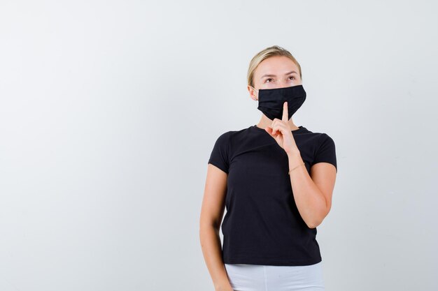 Mujer rubia mostrando gesto de silencio en camiseta negra, pantalón blanco