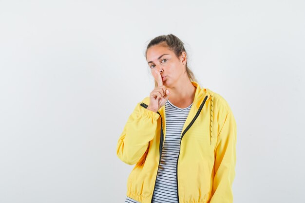 Mujer rubia mostrando gesto de silencio en chaqueta de bombardero amarilla y camisa a rayas y mirando enfocado