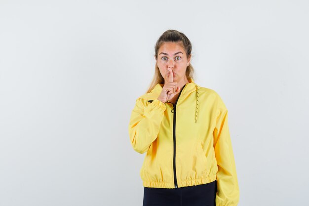Mujer rubia mostrando gesto de silencio en chaqueta de bombardero amarilla y pantalón negro y mirando serio