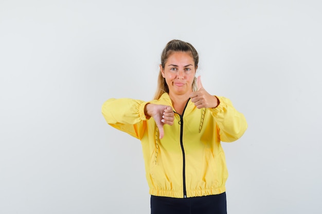 Mujer rubia mostrando el pulgar hacia arriba y hacia abajo en chaqueta de bombardero amarillo y pantalón negro y luciendo bonita