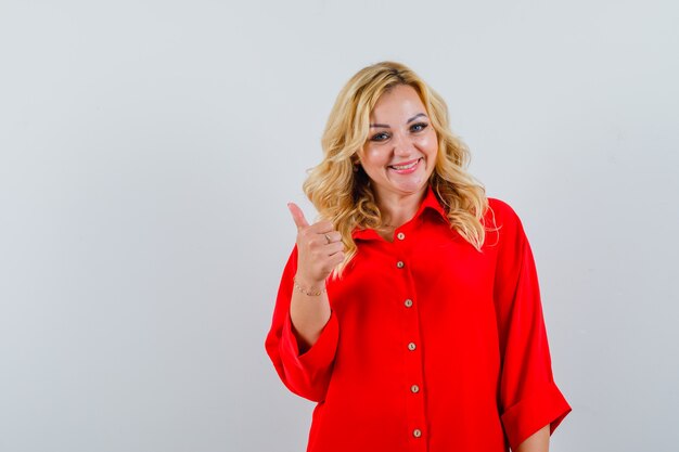 Mujer rubia mostrando el pulgar hacia arriba en blusa roja y mirando feliz.