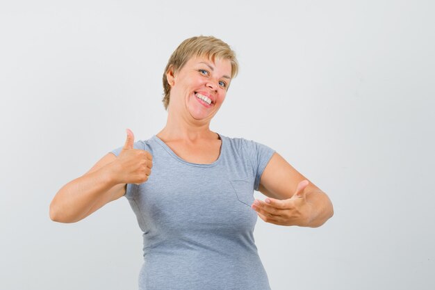 Mujer rubia mostrando el pulgar hacia arriba y pretendiendo sostener algo en su mano con una camiseta azul claro y luciendo alegre. vista frontal.