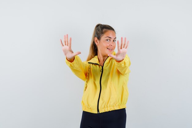 Mujer rubia mostrando señal de stop con ambas manos en chaqueta de bombardero amarillo y pantalón negro y luciendo bonita