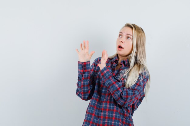 Mujer rubia mostrando señal de stop en camisa a cuadros y mirando sorprendido, vista frontal.