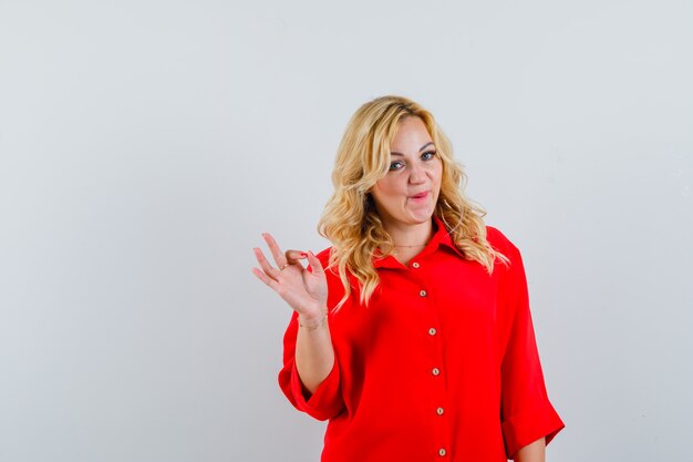 Mujer rubia mostrando signo bien en blusa roja y mirando feliz