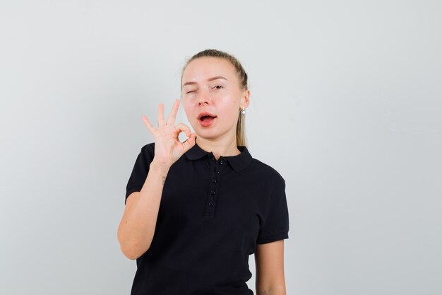 Mujer rubia mostrando signo ok y guiñando un ojo en camiseta negra y luciendo optimista